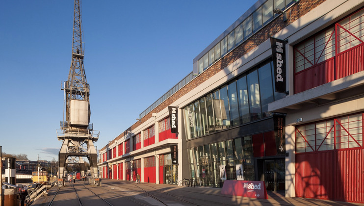 An industrial building with red and white facade, large windows, and a sign reading M Shed on the side is situated near a port area. A large crane is visible to the left of the building, with clear blue skies overhead.