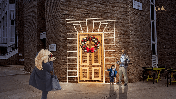 A photographer captures a moment of a man and a young child standing beside a festively decorated doorway with a wreath, illuminated by holiday lights. The scene is set against a brick building in an urban area with street signs visible.