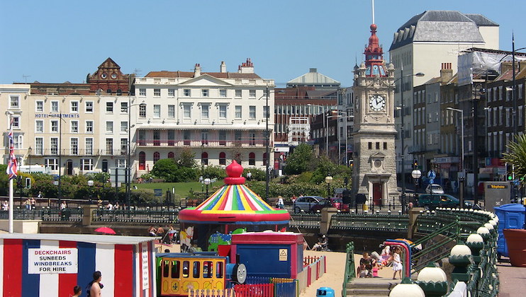 A vibrant seaside scene with a colorful carousel in the foreground and historic buildings in the background. A clock tower is visible to the right, and people are enjoying a sunny day at the beach. Deck chairs and beach huts add to the festive atmosphere.