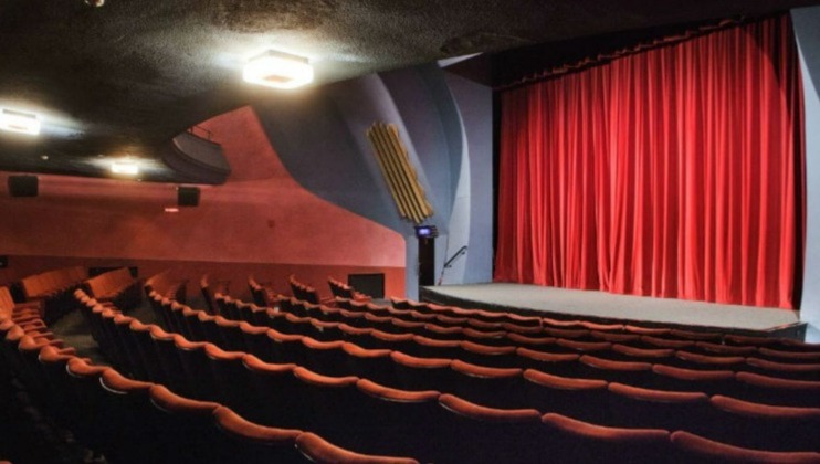 An empty theater with rows of red cushioned seats facing a stage with closed red curtains. The interior is dimly lit, and the walls and ceiling are painted in dark shades. A small staircase and a wooden panel can be seen on the side of the stage.