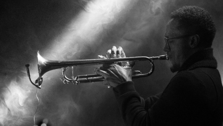 Black and white image of a musician playing a trumpet. The scene is moody with smoke and a spotlight illuminating the musician, emphasizing his focused expression and the shiny surface of the trumpet. The background is dark and hazy.