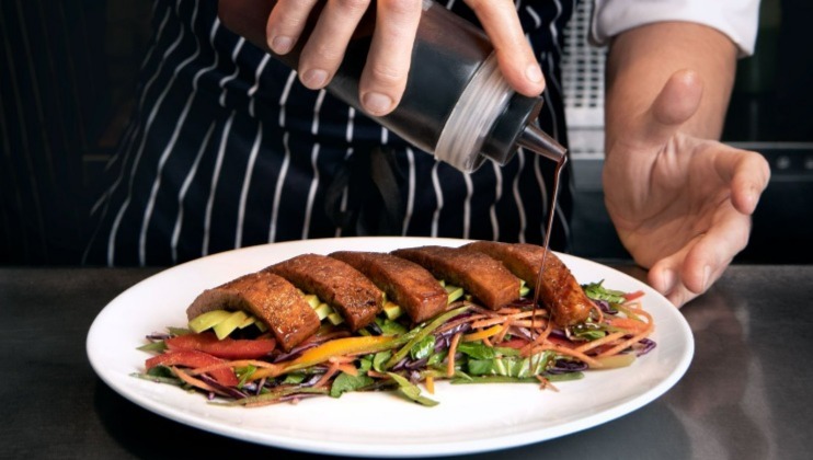 A chef in a striped apron pours sauce over a dish. The plate features several slices of a meat-like entrée, elegantly arranged on a colorful bed of mixed vegetables, including shredded carrots, cucumber, and leafy greens.