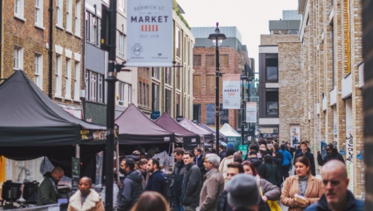 A bustling street market with several people walking and shopping. Various vendors are set up under black and white tents. Signs overhead read Berwick St Market and Market. The street is lined with brick buildings, and the atmosphere is lively and energetic.