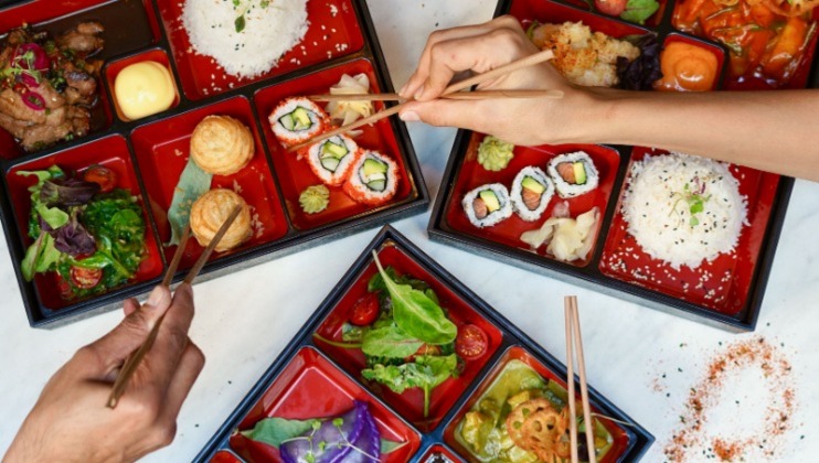 Overhead view of three bento boxes filled with a variety of Japanese dishes, including sushi rolls, rice, salad, and vegetables. Two pairs of hands are using chopsticks to pick food from the boxes. The table is white, and there is a sprinkle of spice in the corner.