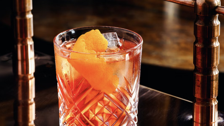A clear, intricately designed glass filled with an amber-colored cocktail, garnished with a twist of orange peel and ice cubes, is placed on a dark, polished surface, with a blurred background featuring copper details.