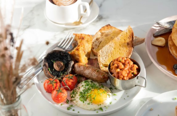 A breakfast plate with toast slices, baked beans in a small cup, a sunny-side-up egg, a sausage, grilled tomatoes, a large mushroom, and a piece of potato or hash brown. In the background, there's a cup of hot chocolate with foam. A dried flower arrangement is on the left.