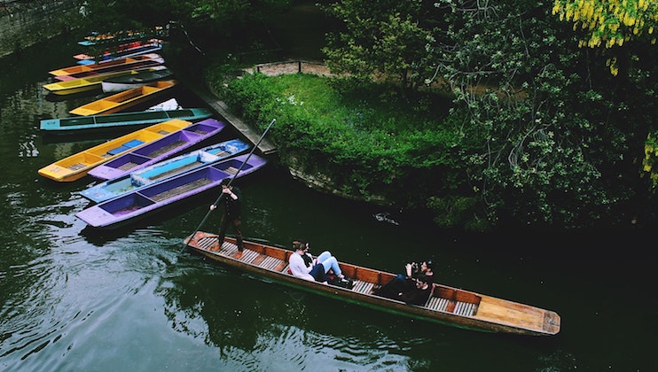 A group of people is punting in a wooden boat along a calm river surrounded by lush greenery. Several colorful flat-bottomed boats are lined up along the bank. The scene is peaceful and serene.