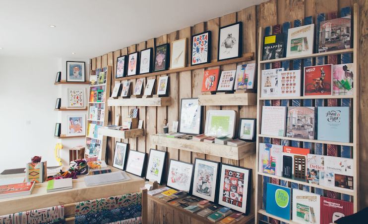 A well-lit art store with various framed artworks, books, and decorative items displayed on wooden shelves along a wall. The shelves are filled with colorful prints, paintings, and graphic designs. A counter with more items is visible on the left side.