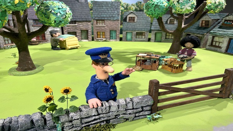A stop-motion animation scene shows a mail carrier in a blue uniform holding a coin by a stone fence. Nearby, a woman stands behind a market stall with items, and another person is carrying a stack of boxes. The backdrop features trees and stone houses.