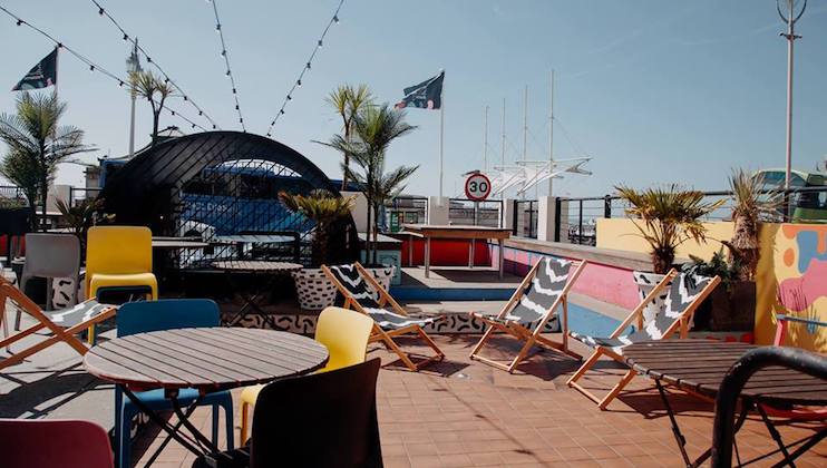 A rooftop patio with several wooden tables and colorful chairs arranged around them. There are also lounge chairs, potted palm trees, string lights, and bright murals decorating the space. A 30 km/h traffic sign is visible in the background. The sky is clear.