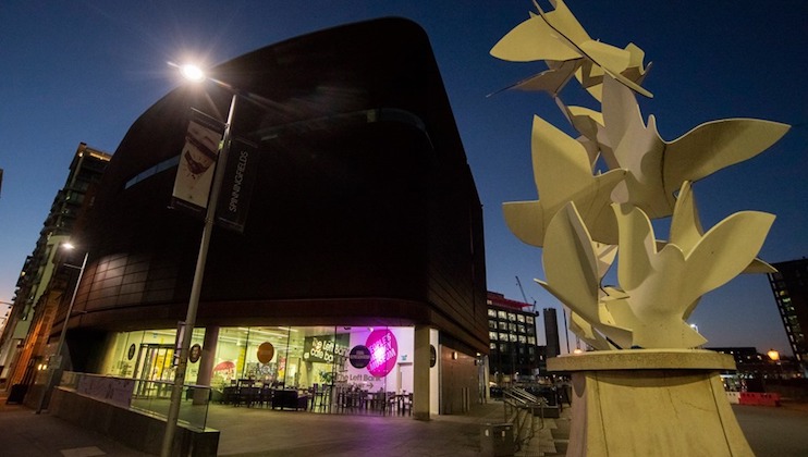 A modern building with a dark exterior and large glass windows is illuminated from the inside at dusk. In the foreground, a white abstract sculpture stands on a pedestal. The sky is dark blue, indicating early evening, and streetlights are lit.