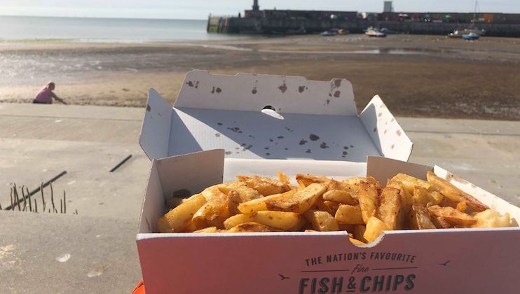 A box of seasoned chips sits on a surface near the shore, with a pier and boats in the background under a clear sky. The text on the box reads, The Nation's Favourite Fish & Chips.
