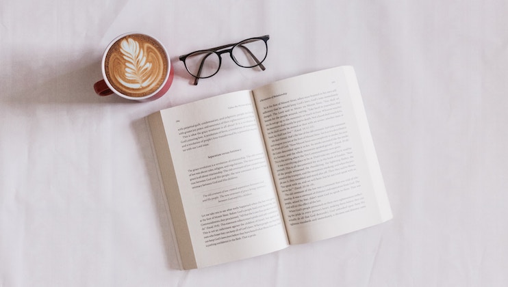 An open book lies on a white surface, with a pair of black eyeglasses placed above it. To the left of the glasses, there is a latte in a brown cup, featuring a leaf pattern created with milk foam.