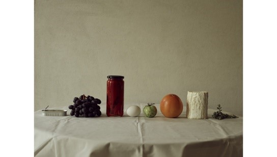 A still life image featuring a variety of objects on a plain table with a white tablecloth. Items from left to right: a small dish, a bunch of grapes, a jar of red preserves, an egg, a green tomato, an orange, a piece of cabbage, and a small sprig of herbs.