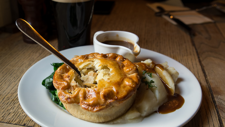 Photograph of a plate of a savoury pie on a bed of mash with gravy and greens