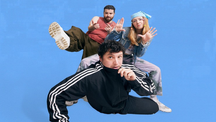 Three young adults strike dynamic, playful poses against a bright blue background. One person crouches in the foreground in an athletic tracksuit, while the other two stand behind, one in casual wear and the other in a denim outfit with a blue bandana.