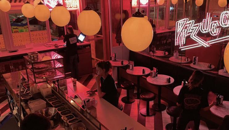 A dimly lit pizza restaurant with neon signs, illuminating the space in red light. Round tables with black chairs are arranged around the room. Two people are working behind the counter, while one person sits at the bar. Large globe lights hang from the ceiling.