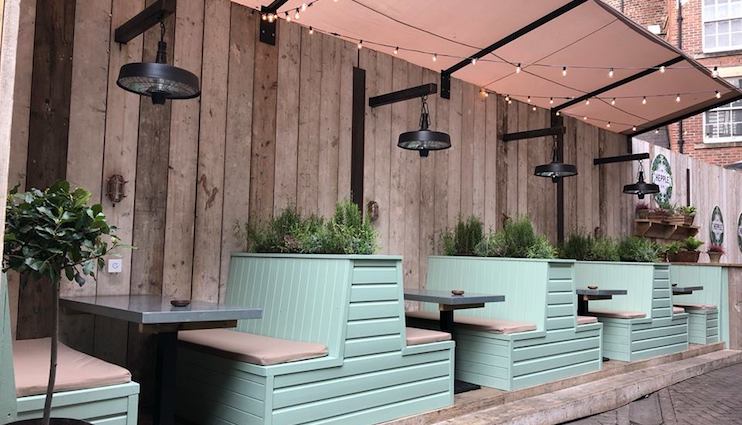 Outdoor seating area with pastel green wooden benches and tables beneath pink awnings. Small potted plants are integrated into the bench backs, and string lights are hung overhead. The area is bordered by a wooden fence and has a cozy, inviting atmosphere.
