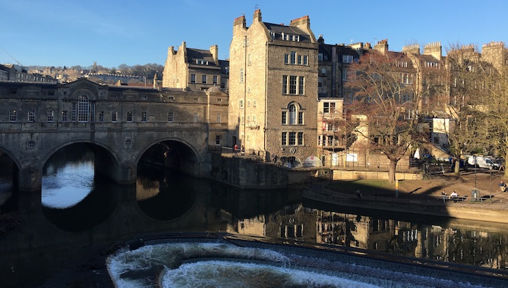 Image of the Pulteney Bridge in Bath, England, with buildings lining both sides of the river. The river has a weir creating cascading waterfalls. The scene is set against a clear blue sky, with some people visible walking and sitting along the riverbank.