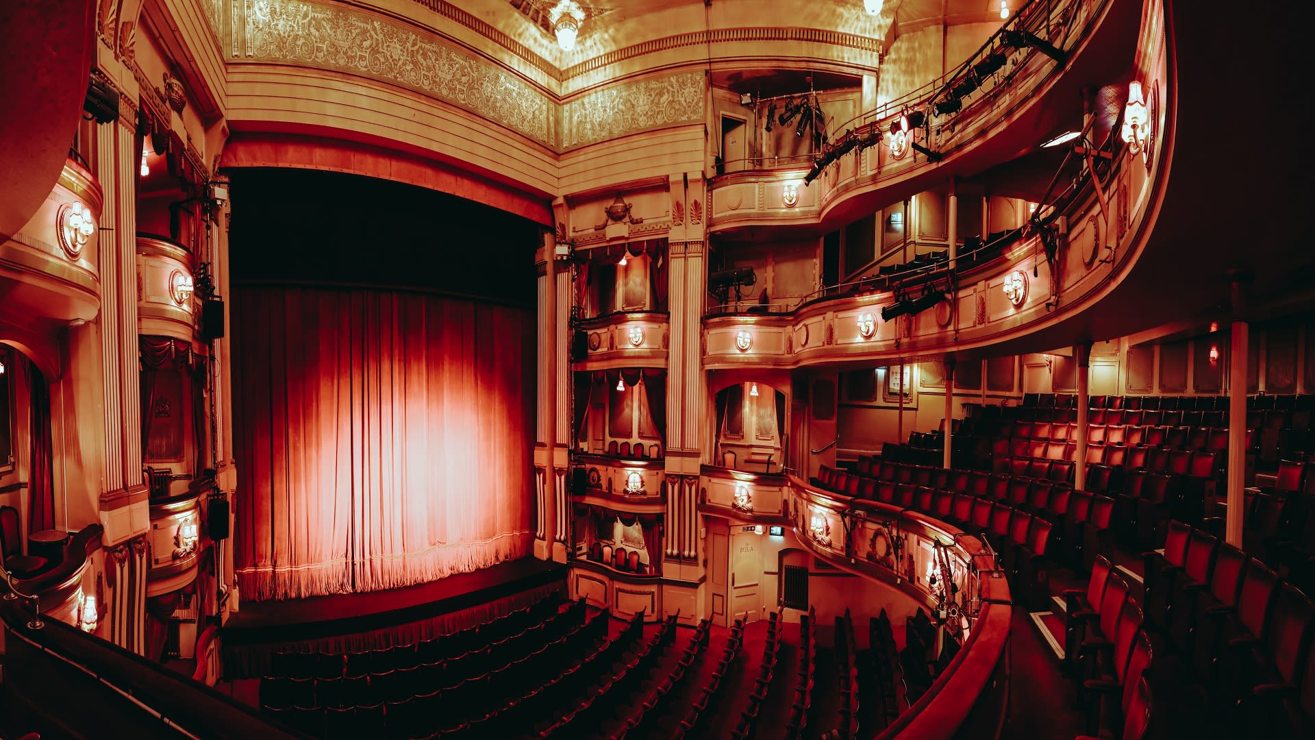 A grand theater with ornate architecture and warm lighting. The red velvet curtain is closed on the stage, and rows of red upholstered seats fill the auditorium. Balconies with additional seating encircle the space, and chandeliers hang from the ceiling.