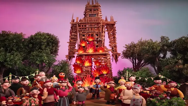 A large wooden structure resembling a human figure is burning, surrounded by trees and a pink sky. In the foreground, a group of claymation-style characters stands together, many dressed in festive attire, with a table of food and decorations nearby.
