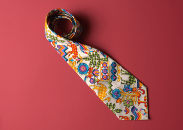 A rolled-up tie lying on a red surface. The tie features a vibrant, multicolored pattern with abstract designs including flowers, zigzags, and geometric shapes in shades of blue, orange, green, and yellow.