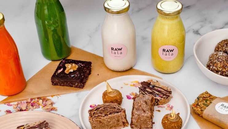 A colorful array of food items and beverages is displayed on a white background. There are green, orange, and yellow juice bottles labeled RAW lala, assorted energy bites, and desserts, including a brownie with a walnut on top and various pastries on plates.