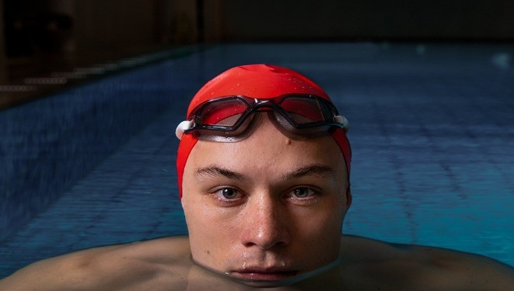 A swimmer with a red swim cap and goggles resting on top of their head is partially submerged in a swimming pool. The swimmer's eyes are focused directly at the camera, with the clear blue water extending into the background.
