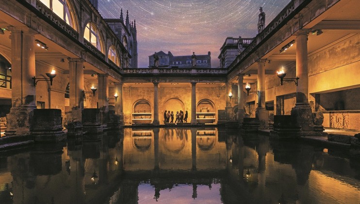 The image captures the Roman Baths in Bath, England, illuminated at night. A group of people stands around the ancient pool, reflecting the warm glow of the torches and the clear night sky filled with stars above. The architecture is grand and historic.