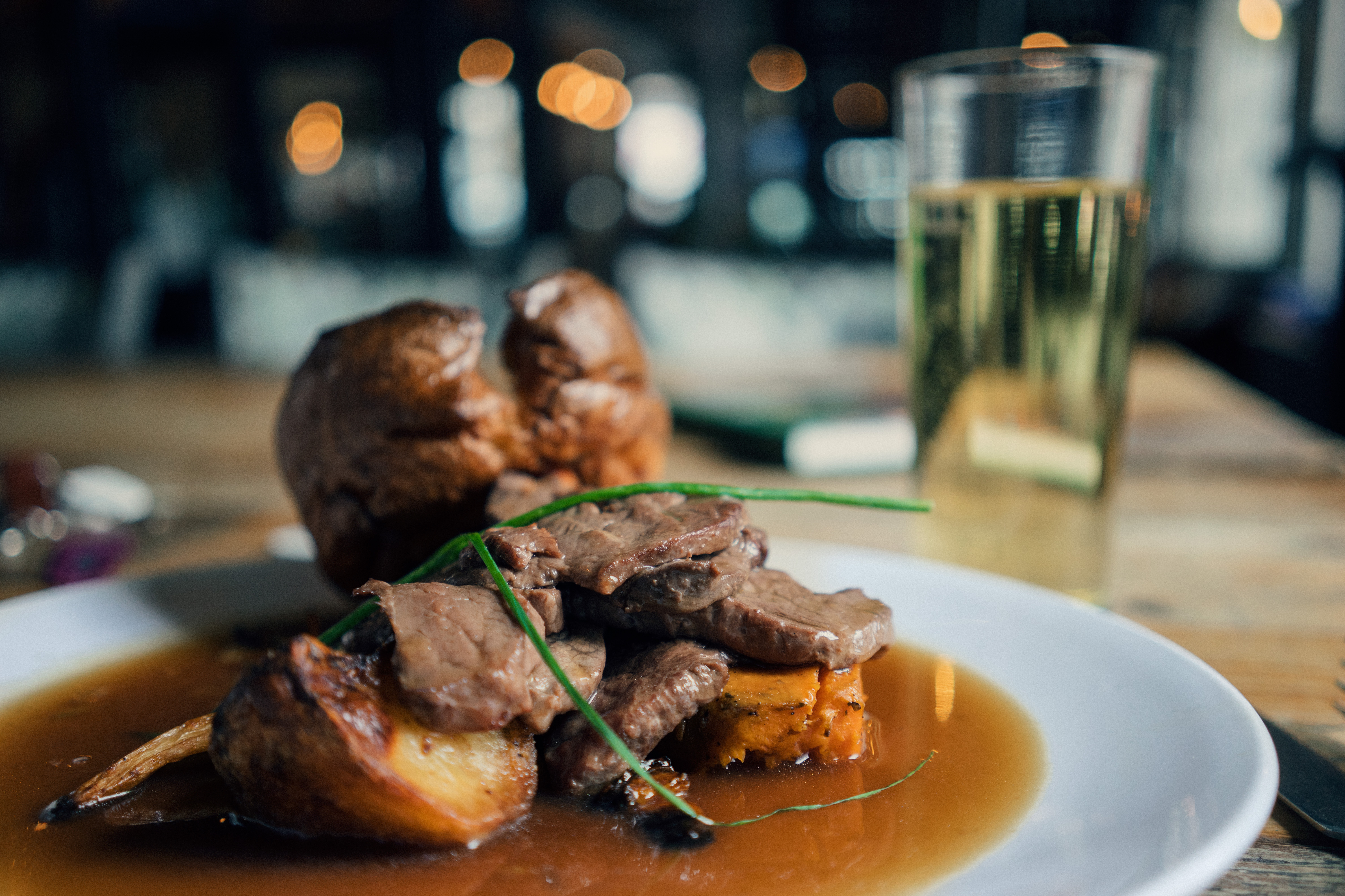 A plate of roast beef slices served with Yorkshire pudding, roasted potatoes, and chives, all drenched in gravy. A glass of light-colored beverage is seen in the background on a wooden table in a dimly lit restaurant with blurred lights.