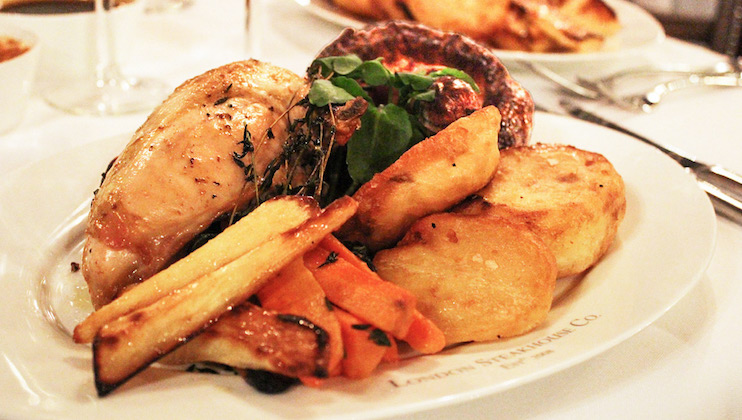 A plate of roasted chicken with crispy skin, served with golden roast potatoes, glazed parsnips, and carrots, garnished with fresh herbs. Another meal is visible in the background. The dish is plated on a white plate with a blurred restaurant setting.