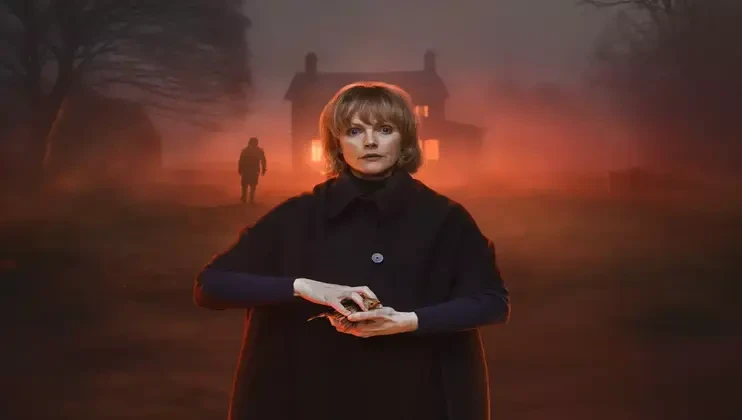 A woman stands in the foreground with a tense expression, her hands together, set against a misty, dimly lit background with an eerie red glow. A shadowy figure approaches from the direction of an old house with glowing windows.