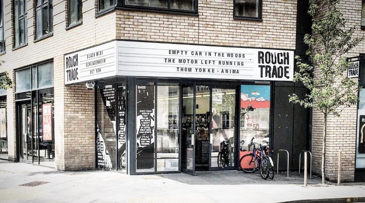 A street corner building with signs for Rough Trade, a music store or venue. The marquee advertises various titles, including Empty Car in the Woods, The Motor Left Running, and Thom Yorke - Anima. A bicycle is parked outside near two small trees.
