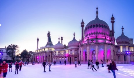People are ice skating at twilight in an open rink in front of the Royal Pavilion, a brightly illuminated historic building with a domed structure and ornate architecture, in Brighton, UK. The sky is clear with a gradient from blue to pinkish hues.