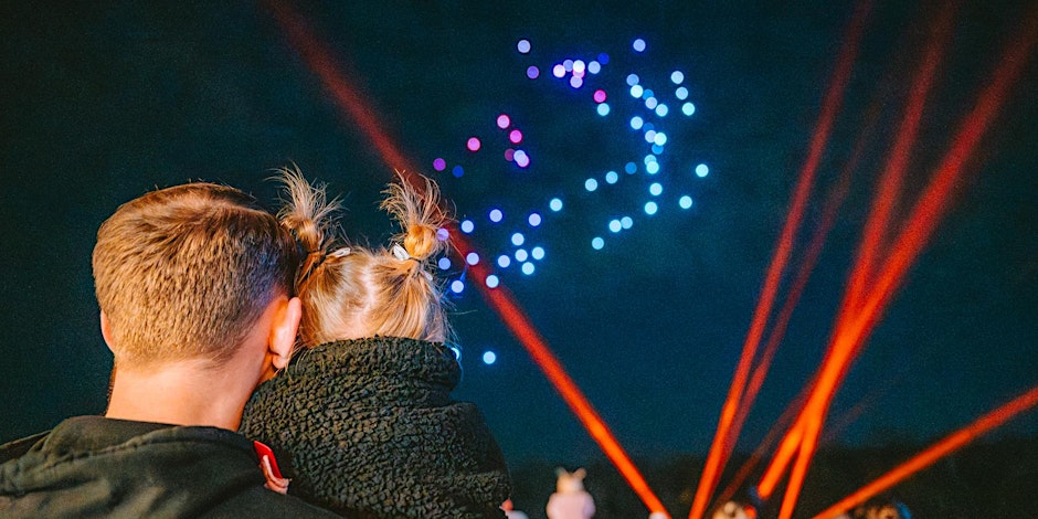 A little girl in pigtails sits on her fathers shoulder wrapped up warm in a teddy coat, watching an overhead Christmas drone light show with lazers.
