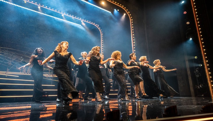 A group of performers dressed in elegant black attire sing and dance in unison on a theater stage with bright lights and dramatic smoke effects. The stage has illuminated steps and curved, light-bordered arches in the background.
