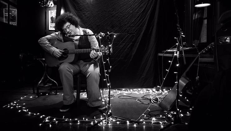 A person sits on a stage playing an acoustic guitar, surrounded by string lights. The room is dimly lit, with a black backdrop behind the performer. The scene has a cozy and intimate atmosphere, enhanced by the warm glow of the lights.