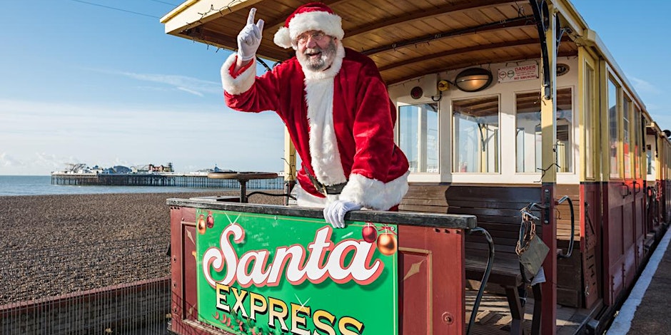 Father Christmas on the Brighton Volks Electric Railway, with the sea and Brighton Pier in the background. He has one finger in the air in a confident, happy expression.