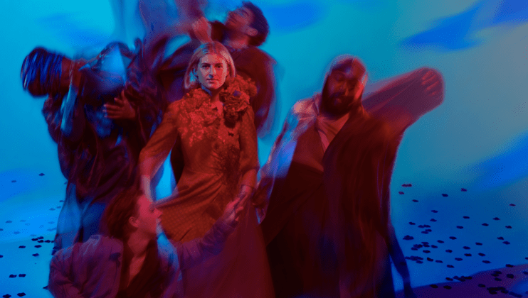 A group of six people dressed in various costumes stand against a blurred, abstract blue and red background. The central woman in a red dress stands out clearly while the others around her are in motion, creating a dynamic and dramatic effect.