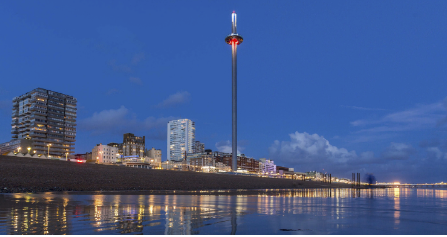 Brighton seafront at dusk, showcasing the illuminated British Airways i360 tower glowing against the blue sky. Tall buildings and apartments line the promenade, their lights reflecting off the calm sea. The pebbled beach stretches along the coastline.