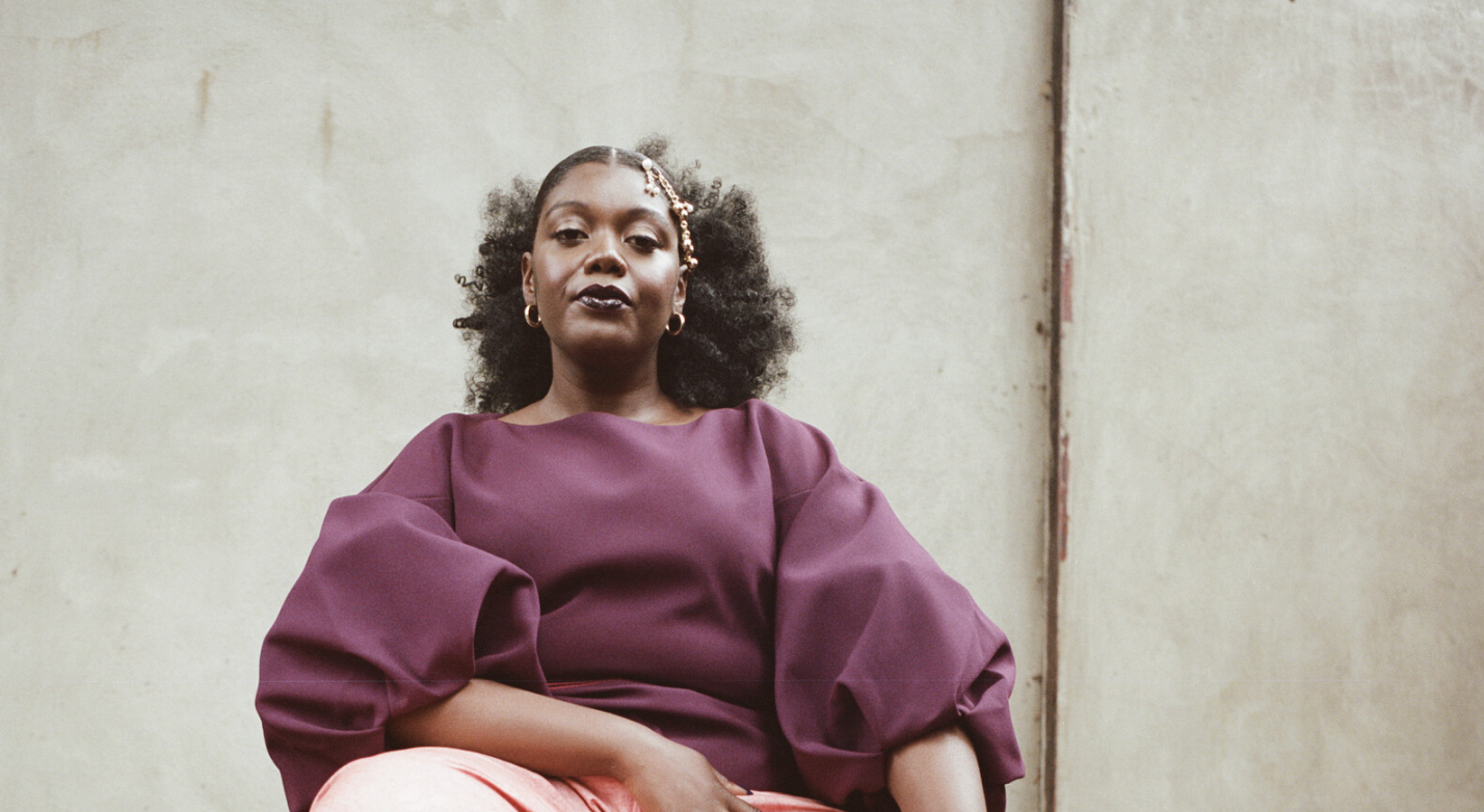 A person with curly hair, wearing a plum-colored top with voluminous sleeves and light pink pants, sits confidently against a textured, neutral-colored wall. They have a calm and poised expression.