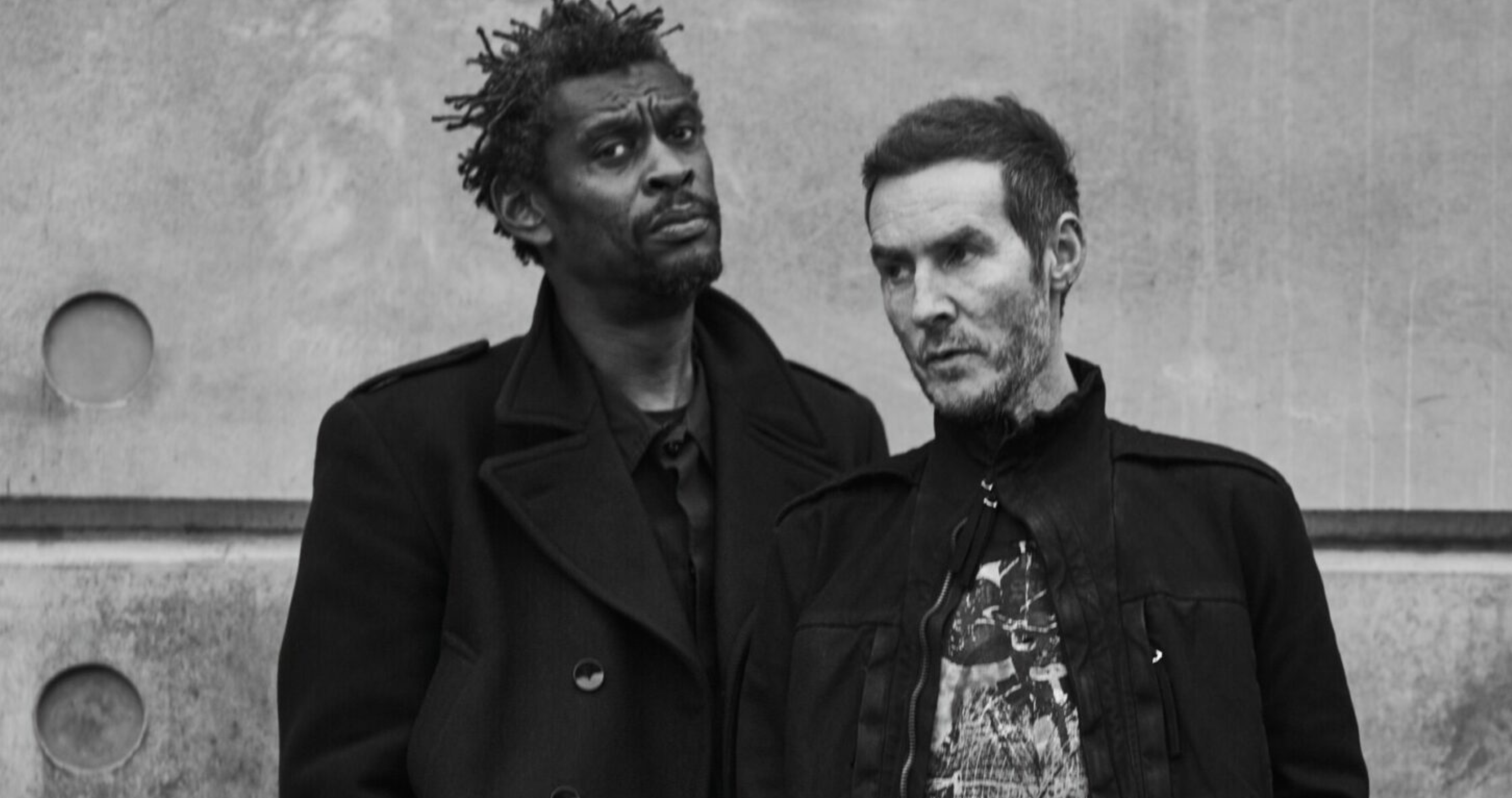 Two men stand side by side against a textured, concrete wall. The man on the left has short, twisted dreadlocks and wears a dark coat. The man on the right has short hair and wears a patterned shirt under a dark jacket. Both have serious expressions.
