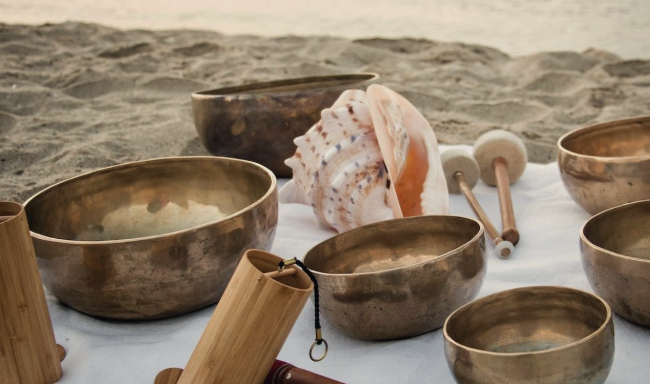 Several brass singing bowls and wooden mallets are arranged on a white cloth on a sandy beach. A large conch shell is placed among the bowls. The setting sun casts a warm light over the scene, creating a peaceful and serene atmosphere.