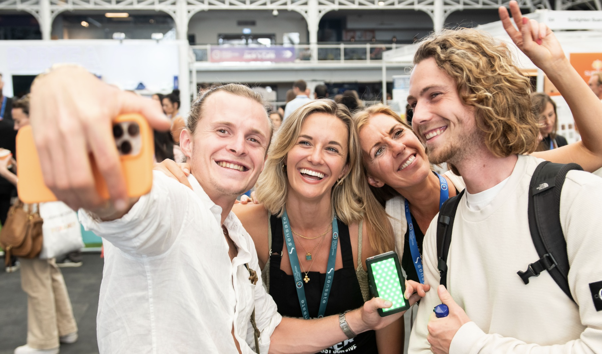 Four people are standing close together, smiling and taking a selfie with a smartphone. They are indoors at what appears to be a busy event or conference, with booths and other attendees visible in the background. Two of them are holding phones.