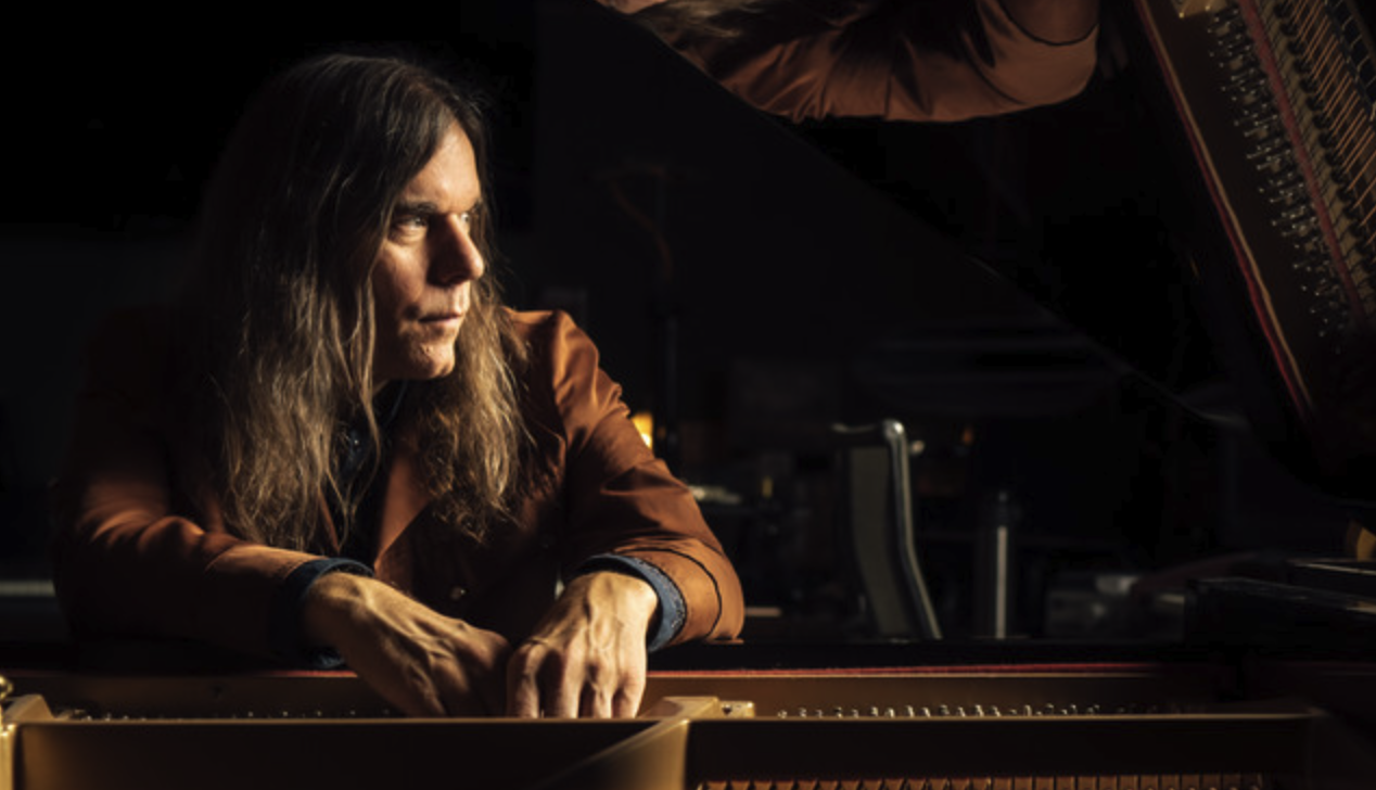 A person with long hair wearing a brown jacket sits at a grand piano, looking thoughtfully to the side. The image is dimly lit, creating a moody atmosphere. The piano's strings are visible, and a reflective surface above shows part of the person's face.