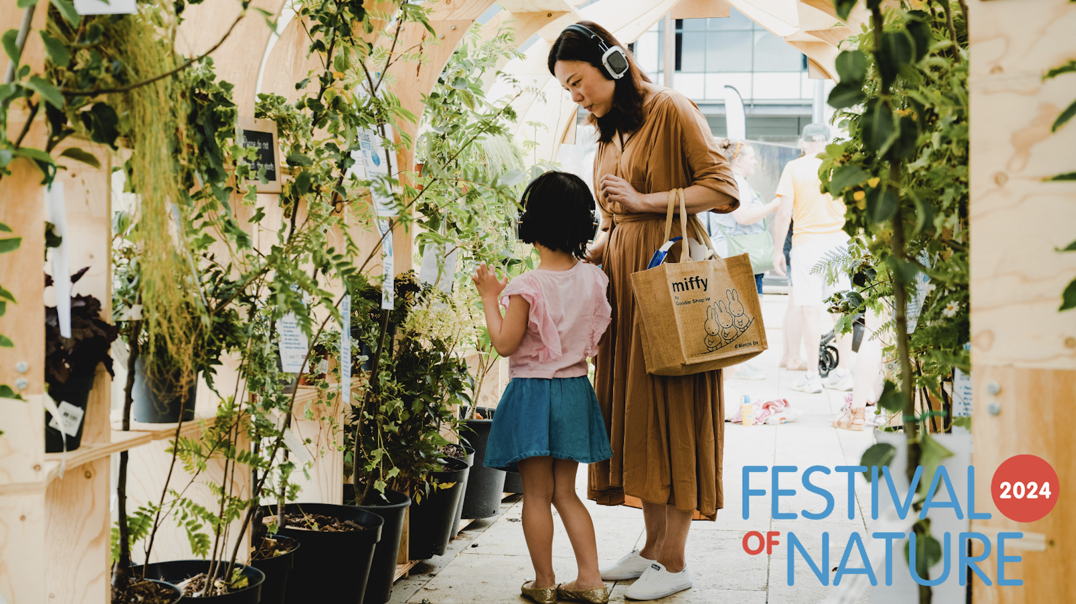 A woman and a child explore plants in a wooden garden structure at the Festival of Nature 2024. The woman wears headphones and carries a shopping bag, while the child, in a blue skirt and pink shirt, looks at a plant. Various potted plants surround them.