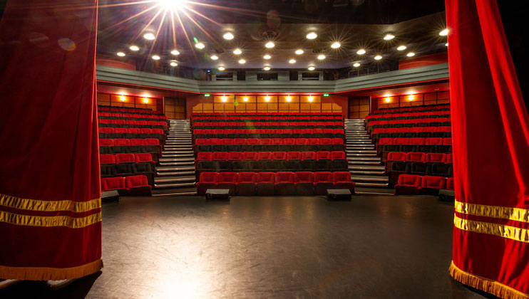A well-lit theater view from the stage, with the audience section filled with red seats arranged in tiers. Bright stage lights shine from above, while two red curtains flank each side of the image.