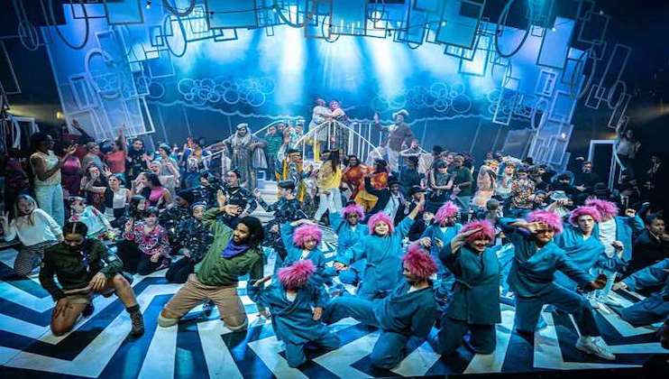 A large, vibrant stage performance features numerous people in eclectic costumes. Performers are dancing energetically under bright blue and white stage lights. The background includes abstract geometric shapes and patterns, adding to the lively and dynamic atmosphere.