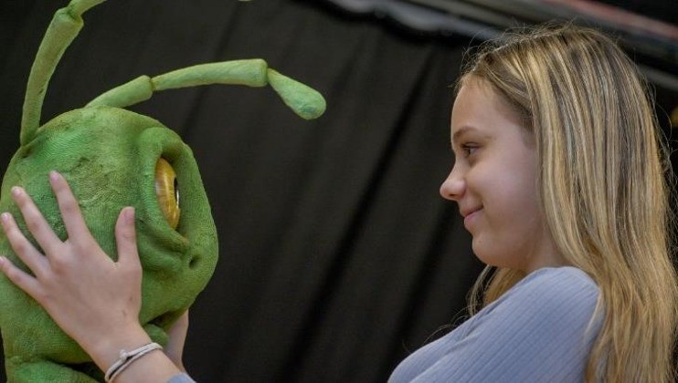 A young girl smiles as she holds the face of a large, green puppet with antennae and a single eye. The background is a dark, out-of-focus curtain.