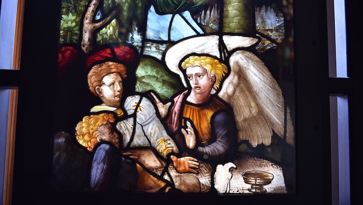 A stained glass window depicts a scene featuring an angel with a white halo attending to a man on the ground. Another figure, dressed in historical attire, looks on with concern. The background shows a natural setting.